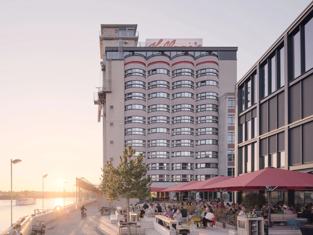 Riverside restaurant terrace with diners under red umbrellas; a tall cylindrical high-rise building along the sunset waterfront.