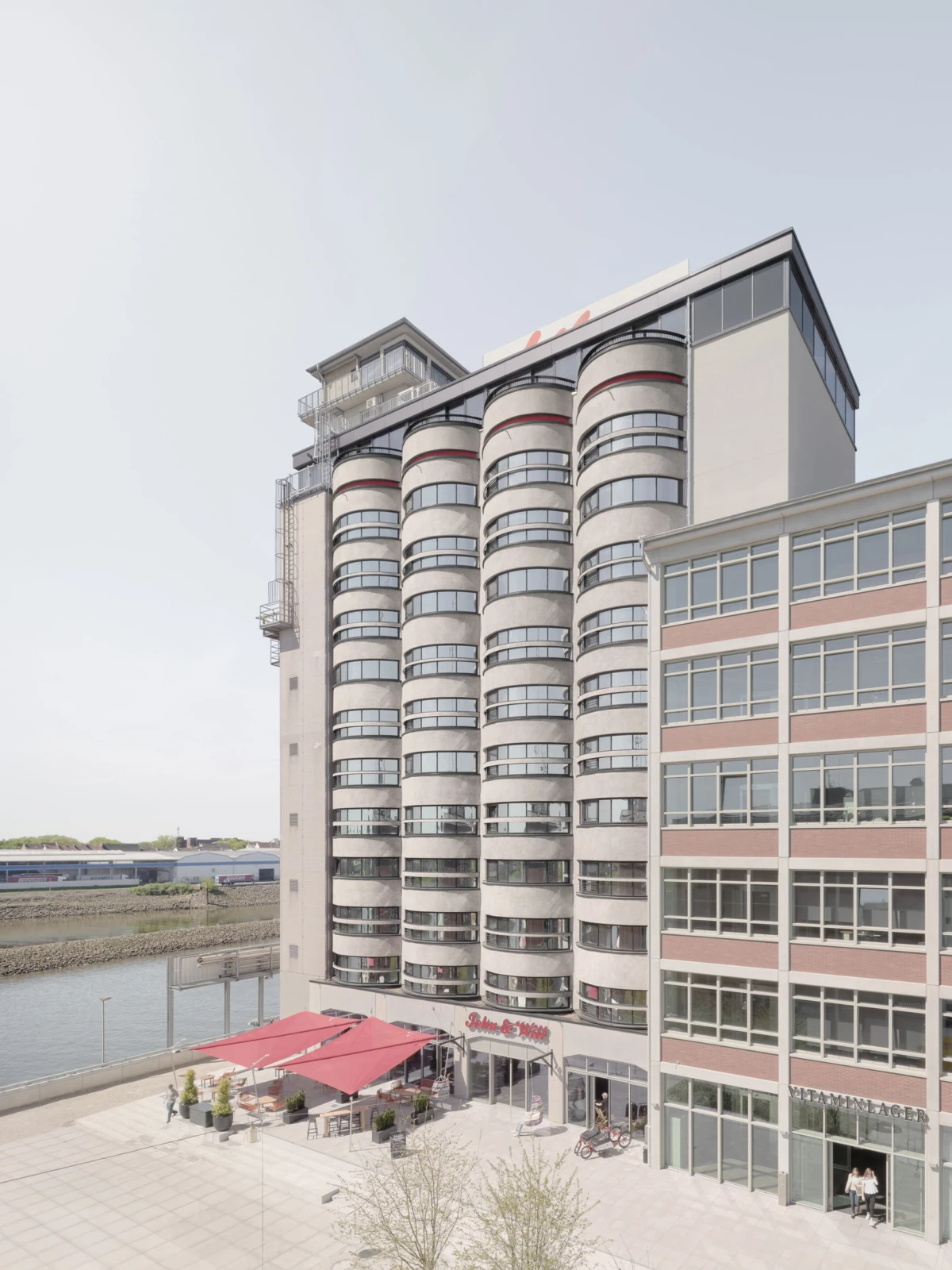 Cylindrical concrete high-rise with rounded balconies beside a waterway; ground-floor cafe patio with red umbrellas and pedestrians.