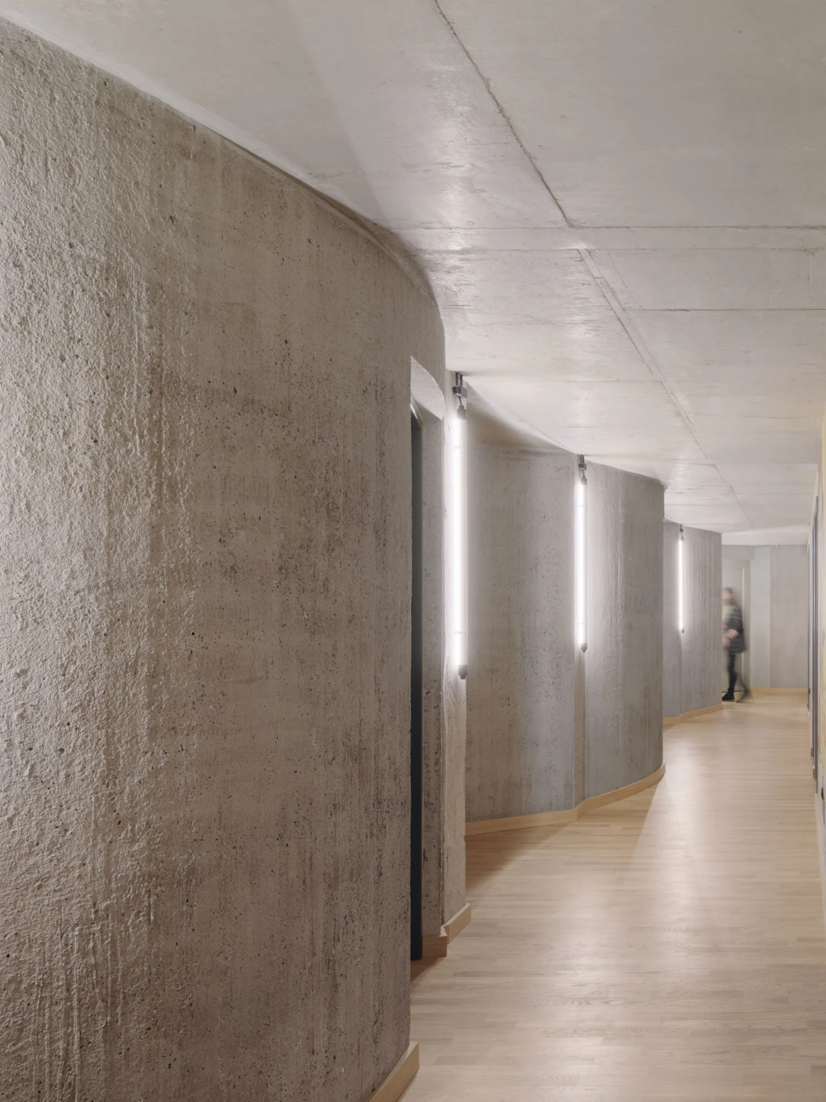 Curved concrete hallway with wooden flooring and wall-mounted vertical light fixtures; a blurred figure walks away in the distance.