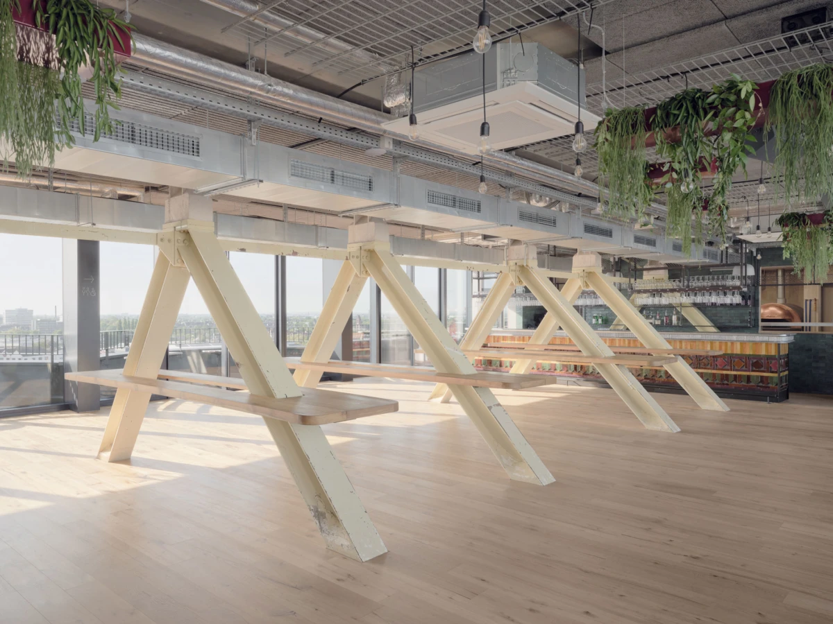 Row of pale yellow A-frame picnic tables in an open, industrial-style space with large windows and hanging plants.