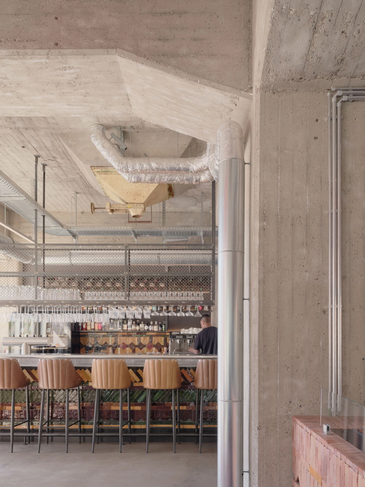 Bartender behind an industrial bar preparing drinks; concrete interior with exposed pipes and tan high-back stools.