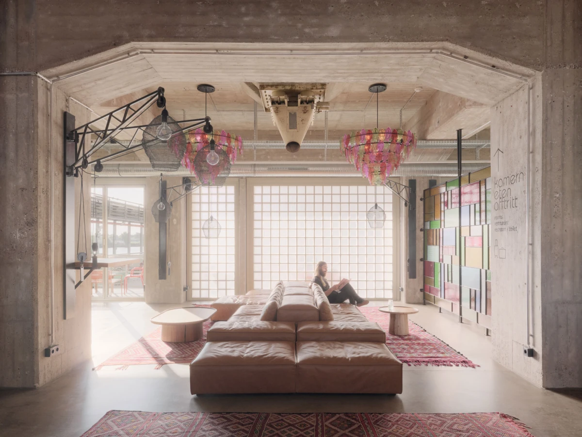 A person sits on a tan sectional sofa in a bright industrial loft, reading a book amid modern hanging lights and colorful wall panels.