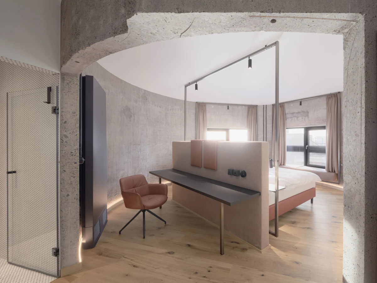 Hotel room with curved concrete walls, a partitioned desk, pink upholstered chair, and a bed beside large windows.
