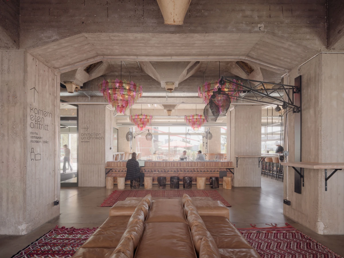 Person seated at wooden counter in an industrial-style cafe; leather sofas, red patterned rugs, and colorful hanging lanterns.