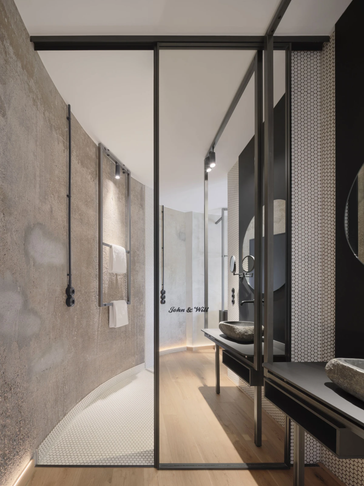 Modern bathroom interior with curved concrete walls, glass shower enclosure, light wood floor, black metal frames, and double stone vessel sinks.