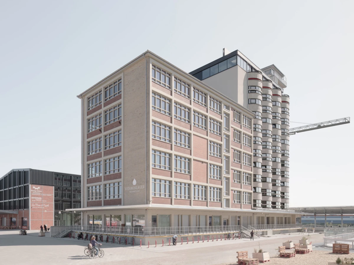 Brick-and-glass five-story building with a cylindrical residential tower on the right; construction crane nearby; pedestrians and cyclists in the foreground.