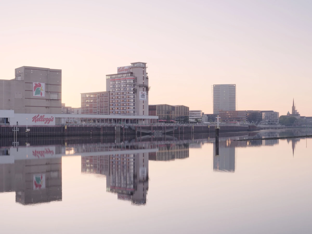 Riverside city skyline at dawn with Kellogg's building; calm water reflects the scene.