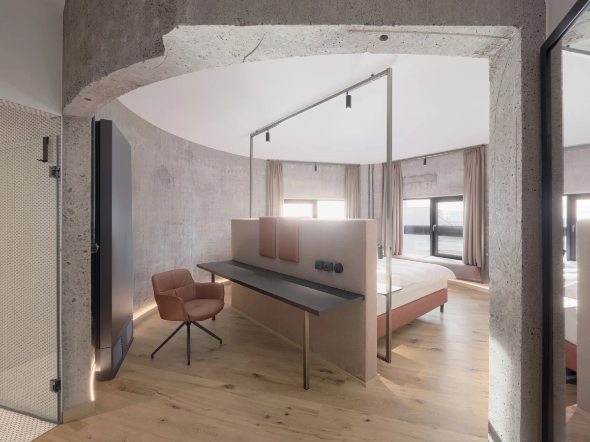 Modern hotel bedroom with curved concrete walls, a bed, built-in desk, pink upholstered chair, and large windows with beige curtains.
