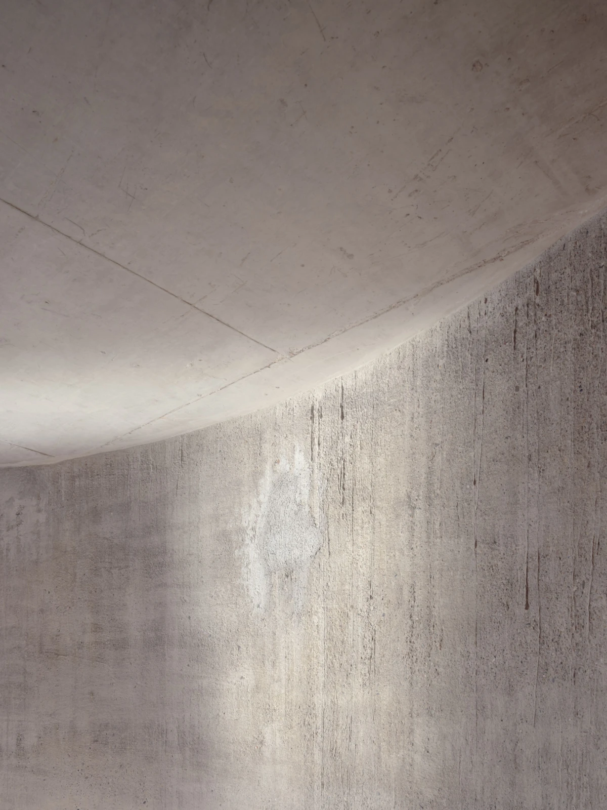 Curved concrete ceiling meets a streaked concrete wall; gray interior with vertical stains and soft shadows.