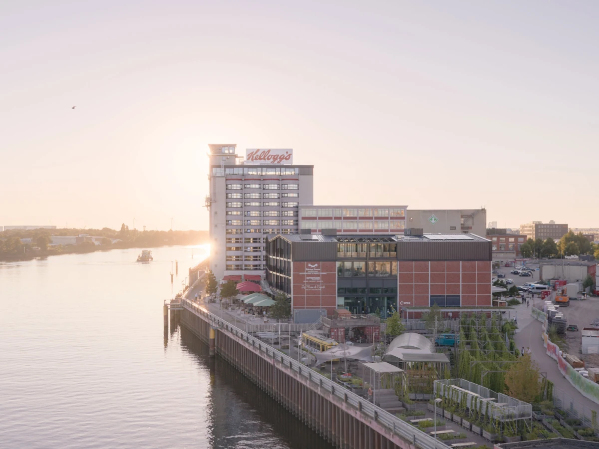 Riverside complex at sunset with a tall white building bearing Kellogg's sign beside a brick-and-glass office block and a boardwalk.
