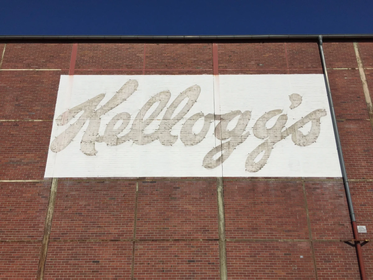 Kellogg's script logo painted on a white panel on a brick wall.