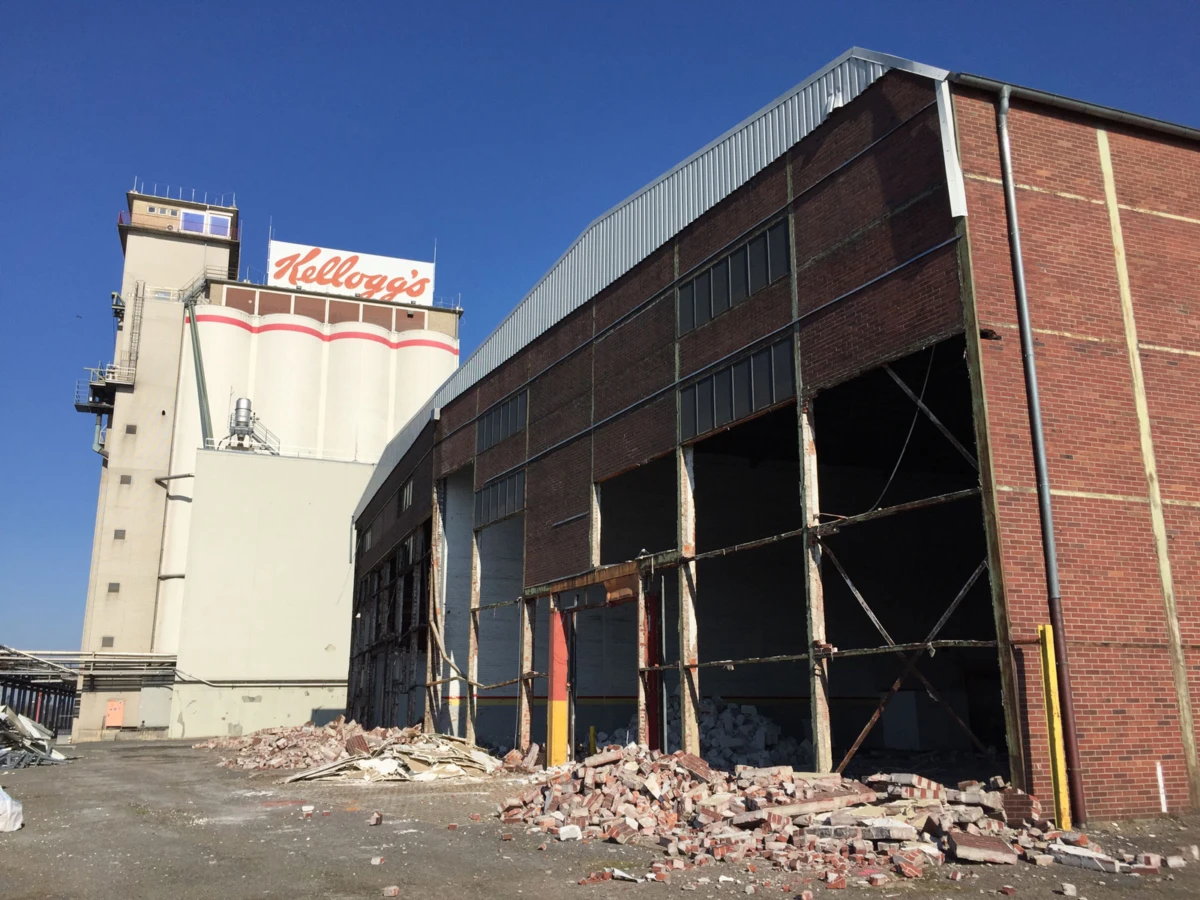 Brick industrial warehouse under demolition; piles of bricks in the foreground, tall silo with Kellogg's sign in the background under a blue sky.