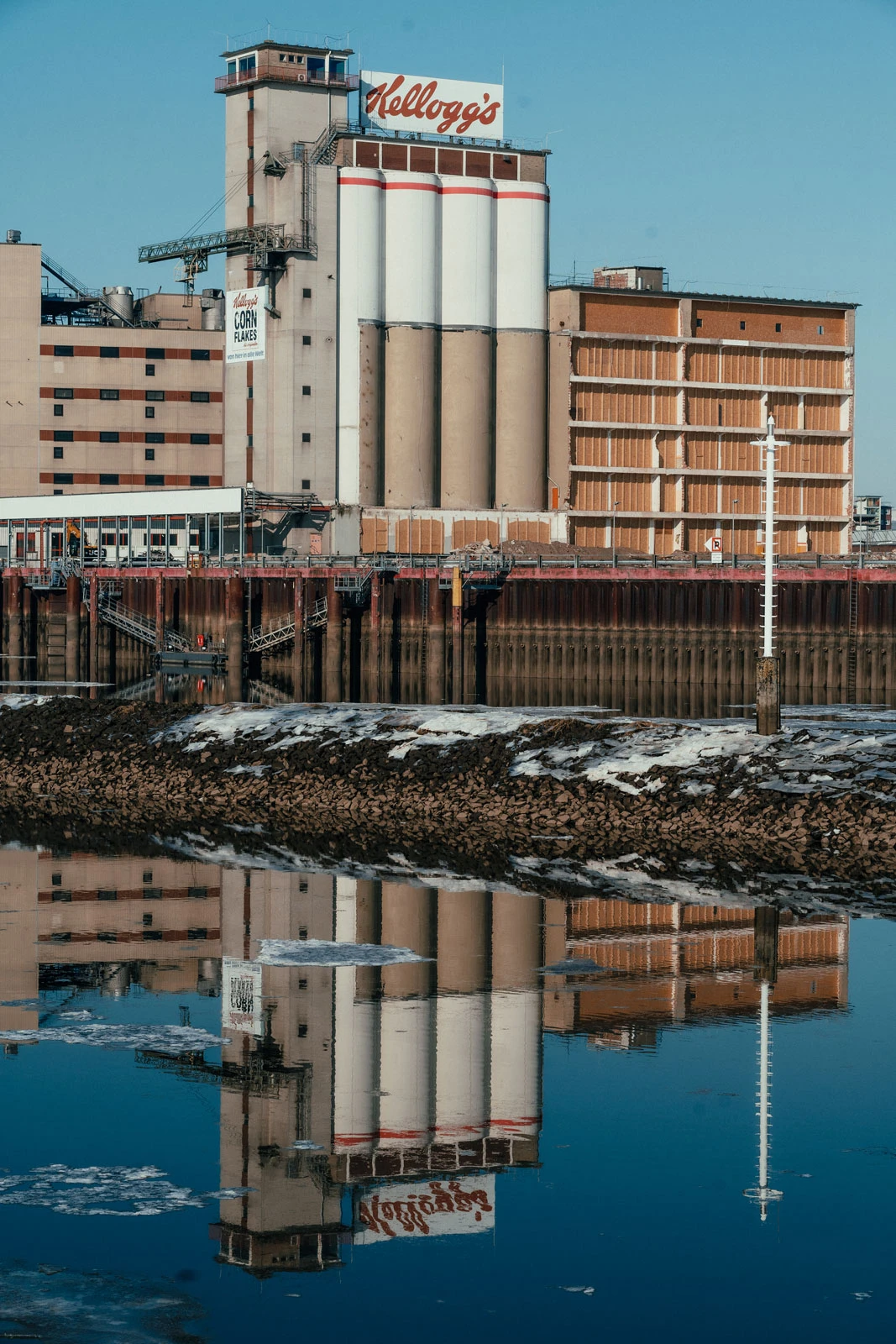 Kellogg's cereal factory with tall silos on a waterfront, reflected in the calm river.
