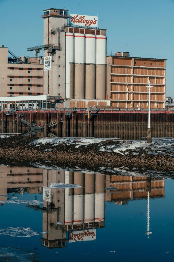 Kellogg's grain silo complex by a harbor; tall white silos and brick buildings with a dock, reflected in calm water.