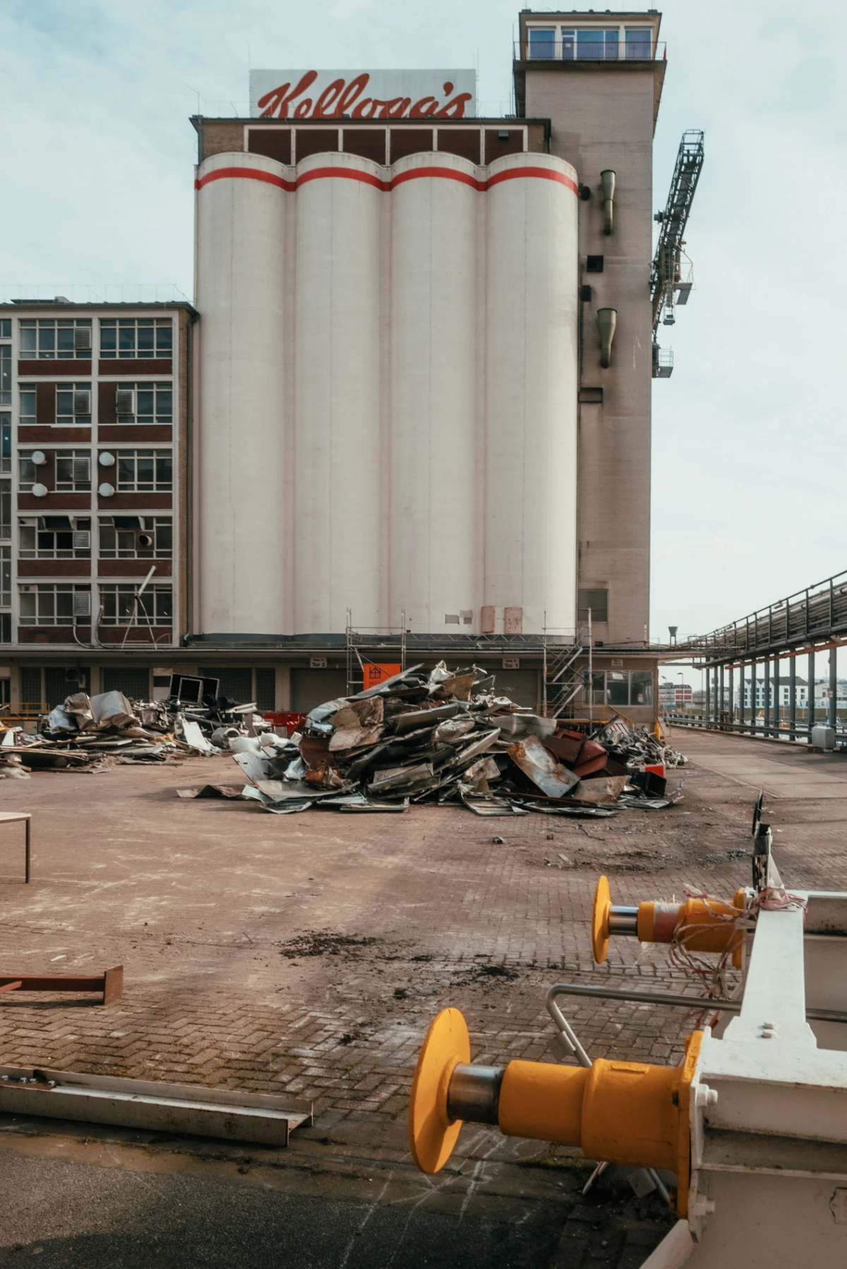 Industrial facility with tall white silos and brick building; Kellogg's sign; pile of scrap metal on pavement and yellow cable reels in foreground.