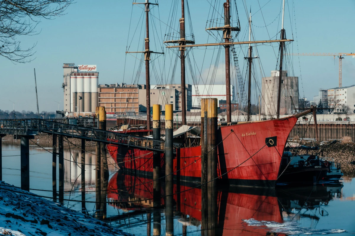 Red sailing ship with tall masts moored at a harbor; calm water reflects the vessel and industrial skyline.