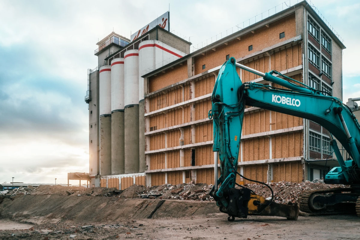 Blue hydraulic excavator at a demolition site beside a partially stripped brick building and concrete silos, rubble on the ground.
