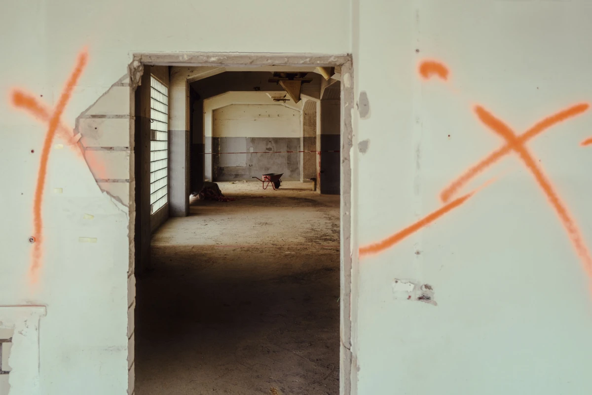 Abandoned building interior; doorway opens to a long, dim hallway with a wheelbarrow and debris, orange X graffiti on pale walls.