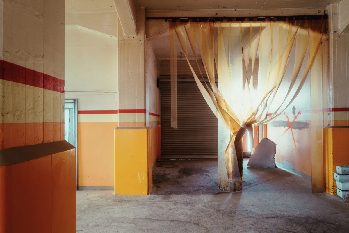 Industrial corridor with orange walls and red stripe; central fabric strips tied into a tent-like curtain in front of a closed metal roll-up door.