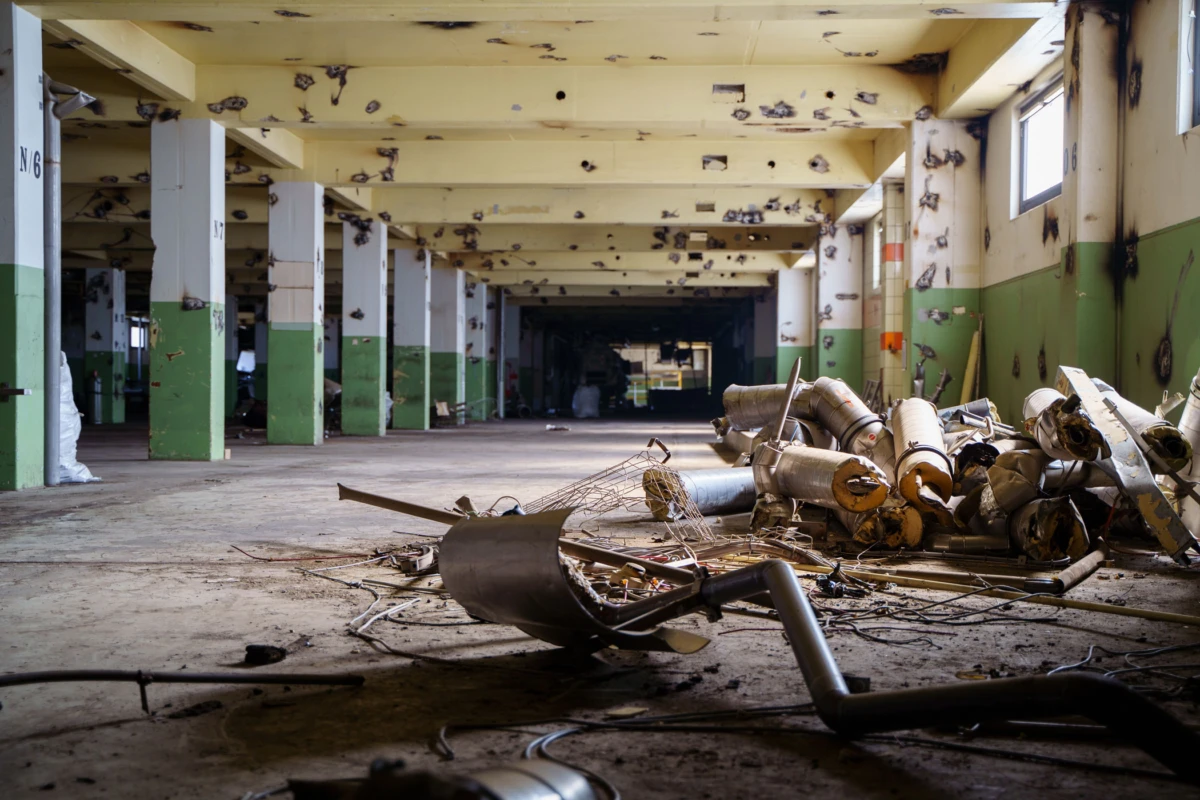 Abandoned industrial warehouse interior with green-walled columns, scattered rusted pipes and debris on a concrete floor.