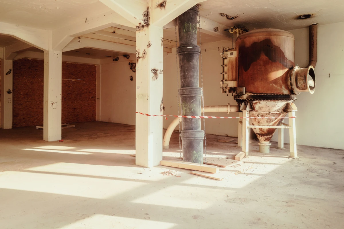 Sunlit industrial room with white columns and a large rusted cyclone dust collector connected to pipes, cordoned off by red-and-white tape.