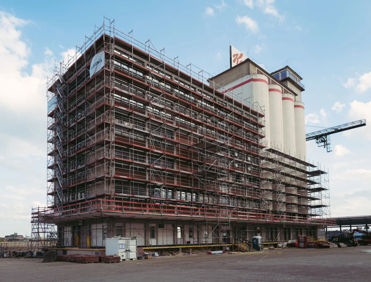 Multistory industrial building under construction wrapped in extensive scaffolding; cement silos in the background with a crane overhead.