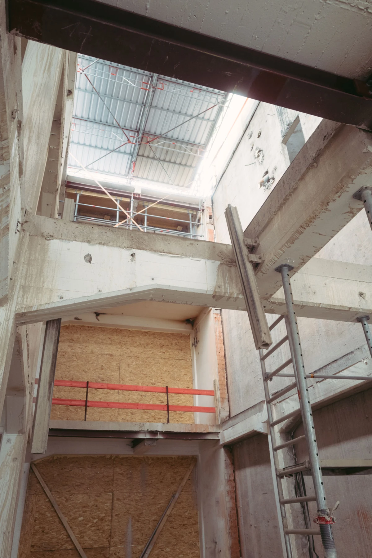 Unfinished concrete building interior with metal scaffolding, a ladder, and plywood walls; open atrium with skylight.