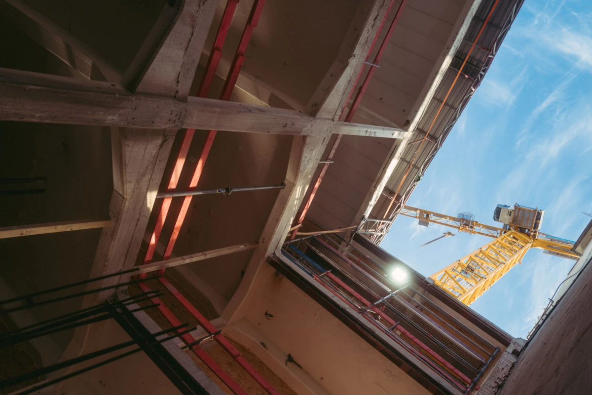 Interior view of a multistory construction site with exposed wooden beams and red pipes; a yellow tower crane rises against a blue sky.