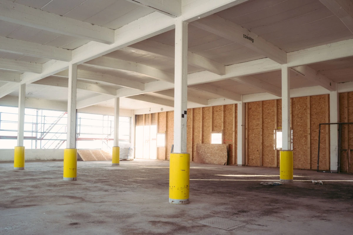 Empty warehouse interior with white columns, yellow protective padding, plywood walls, and scattered construction materials.