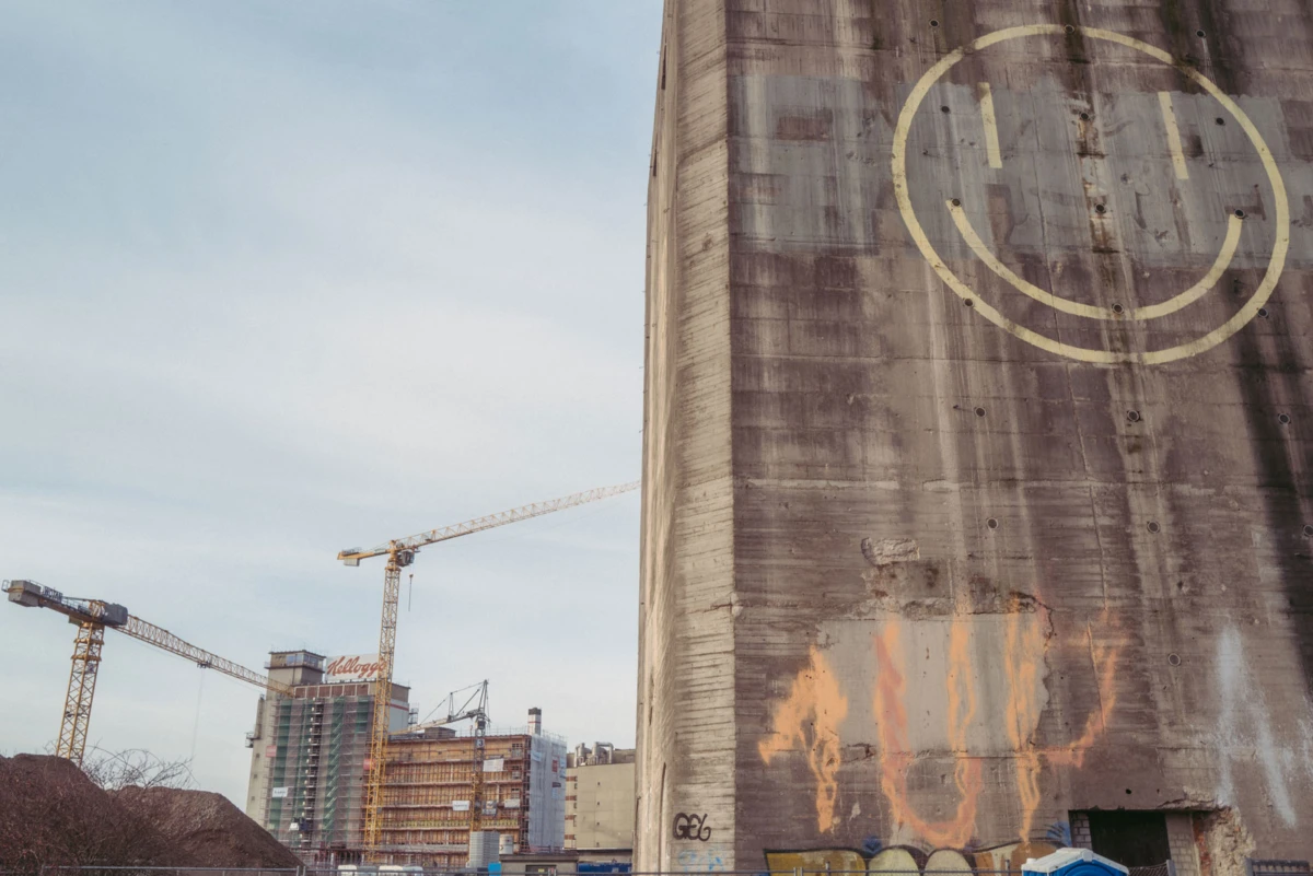 Concrete silo with a large smiley-face graffiti; construction site with cranes and buildings in the background.