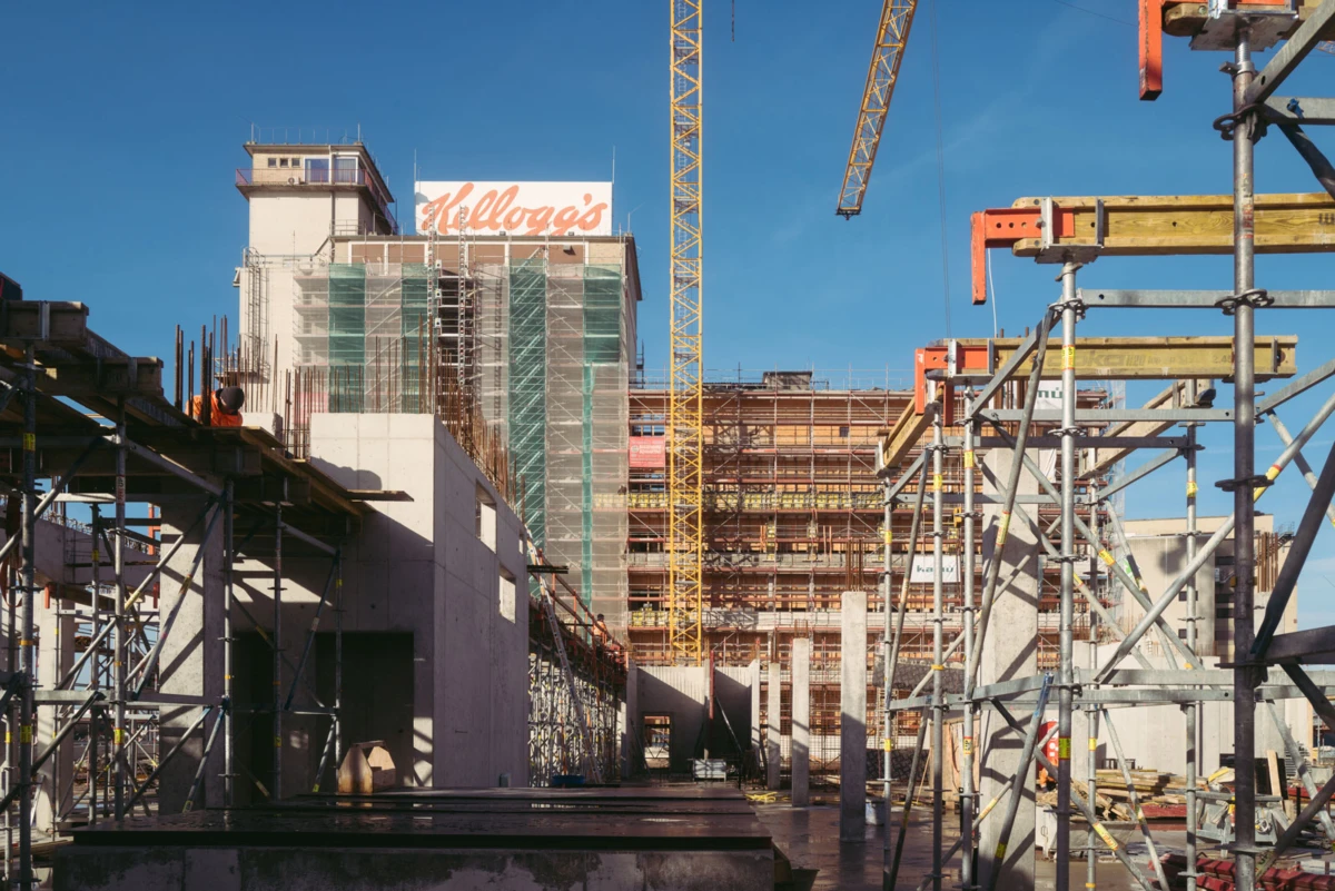 Construction site with steel scaffolding, concrete structures, and a tower crane beside a multi-story building under construction.