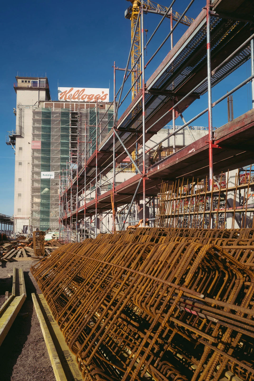 Construction site with rusted rebar cages in the foreground, scaffolding, and a crane beside a multi-story building under construction.