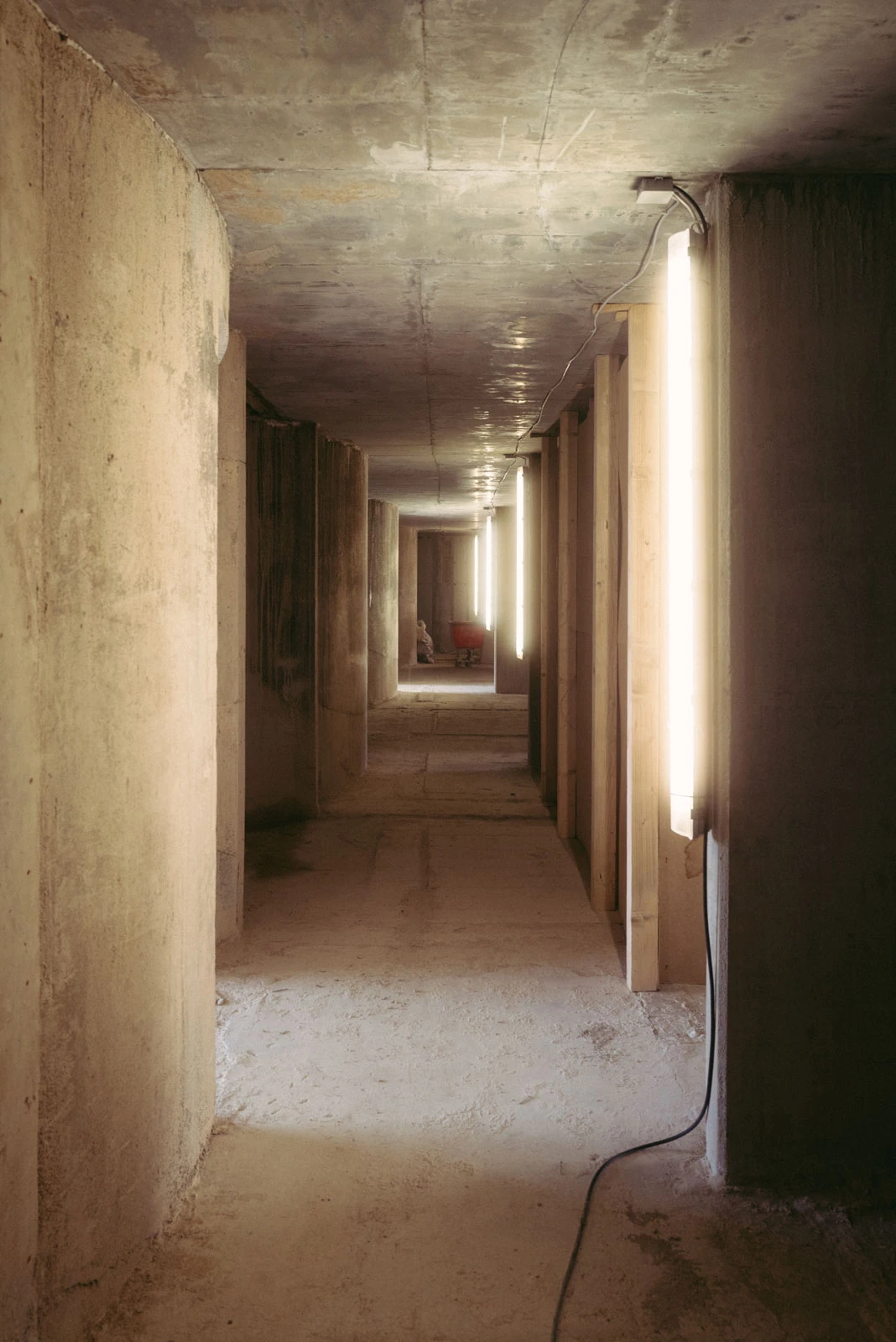 Concrete construction-site hallway with evenly spaced columns and vertical light tubes; wheelbarrow visible at the far end.