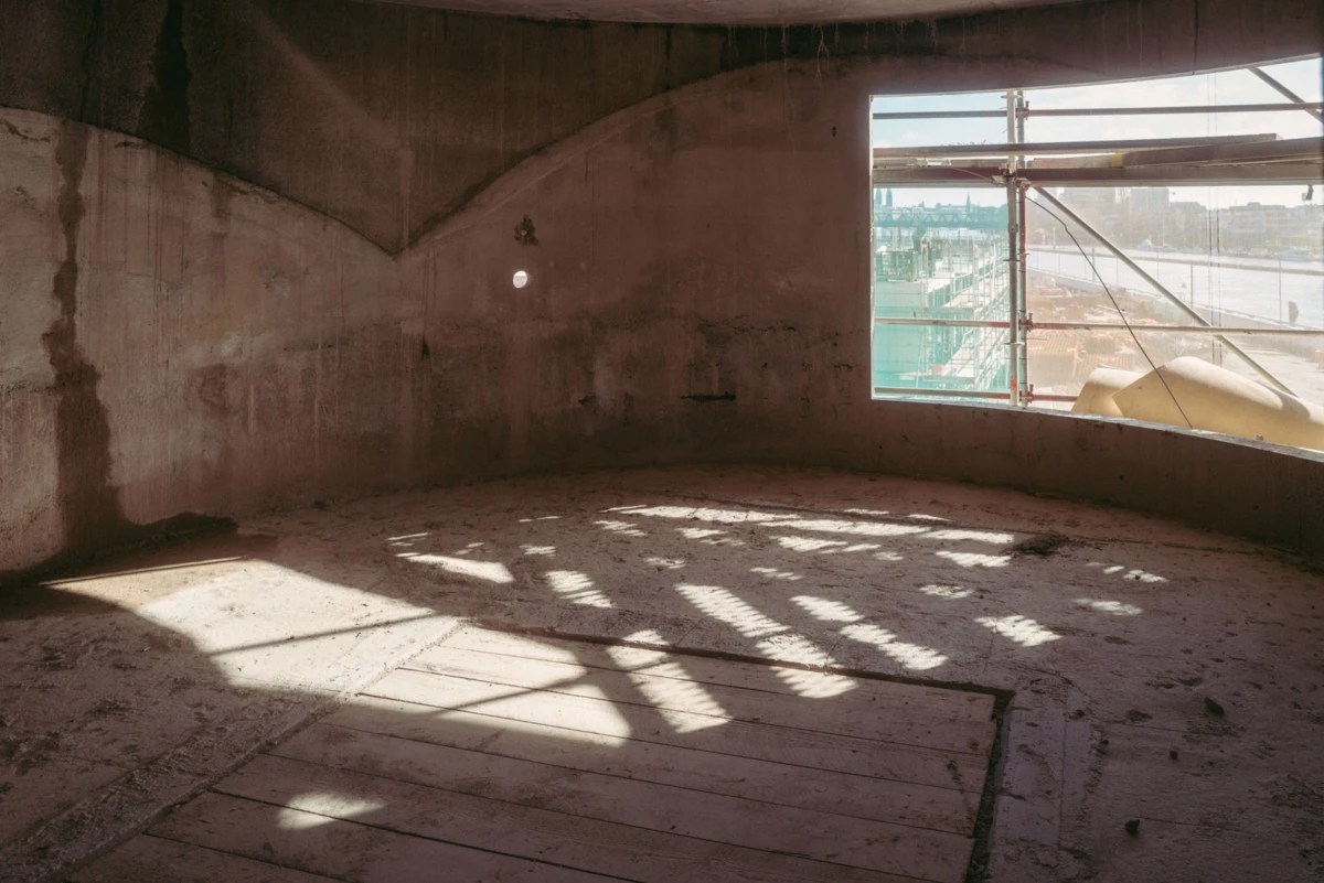 Unfinished circular concrete interior under construction; scaffolding visible through a window, sunlight casting geometric shadows on the floor.