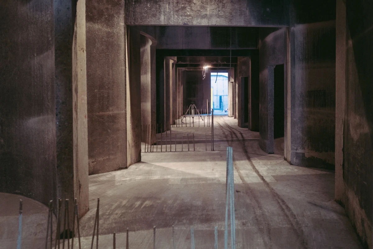 Concrete construction corridor with large pillars and exposed rebar, leading to a blue-lit doorway at the far end.