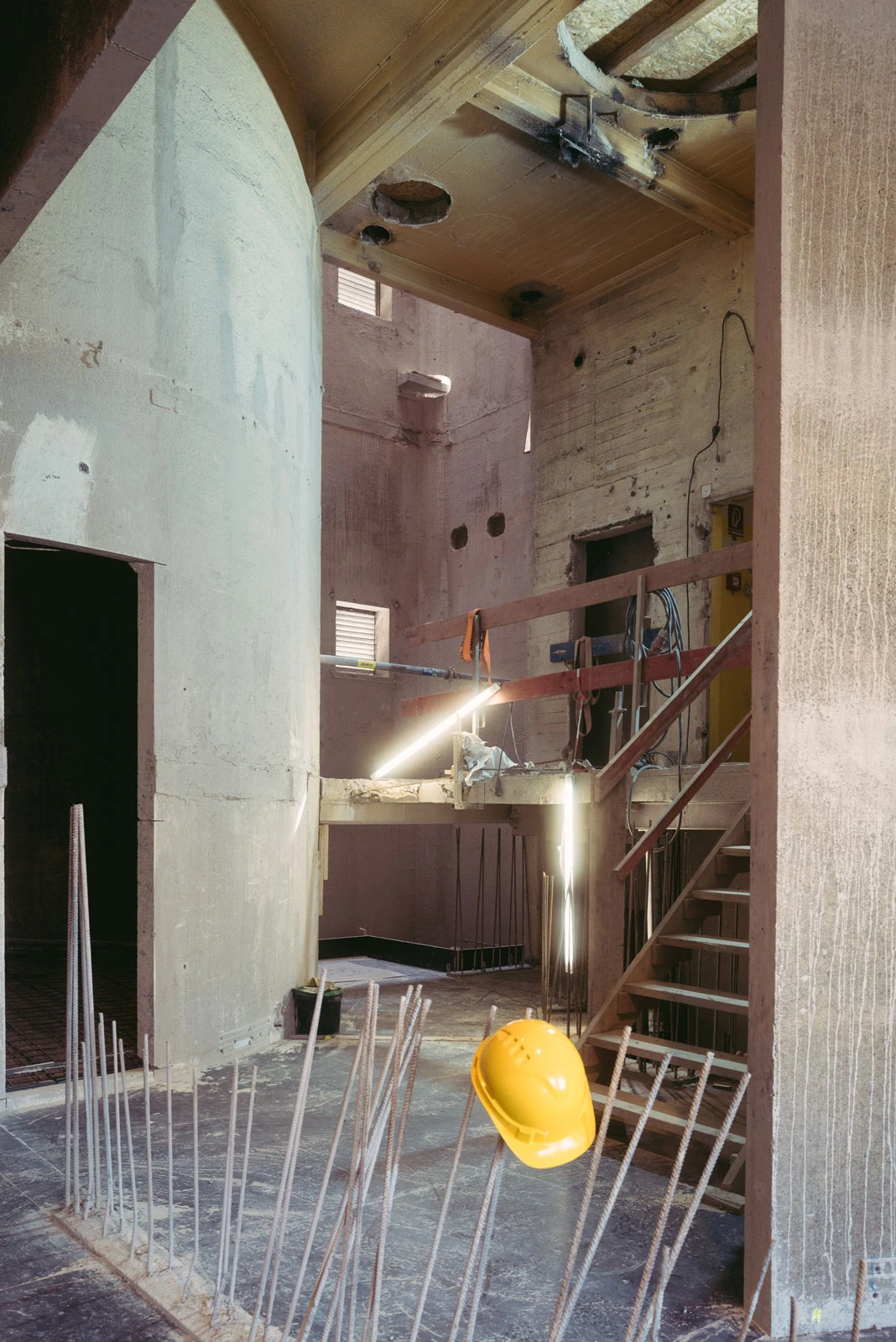 Construction site interior with exposed rebar, concrete columns, and a suspended yellow hard hat.