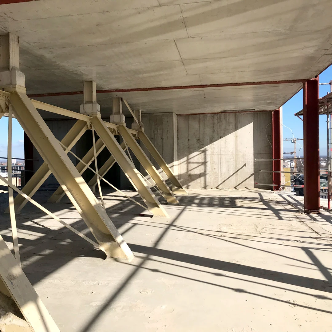 Beige diagonal steel braces support a concrete slab at a construction site; red vertical columns and city view in distance.