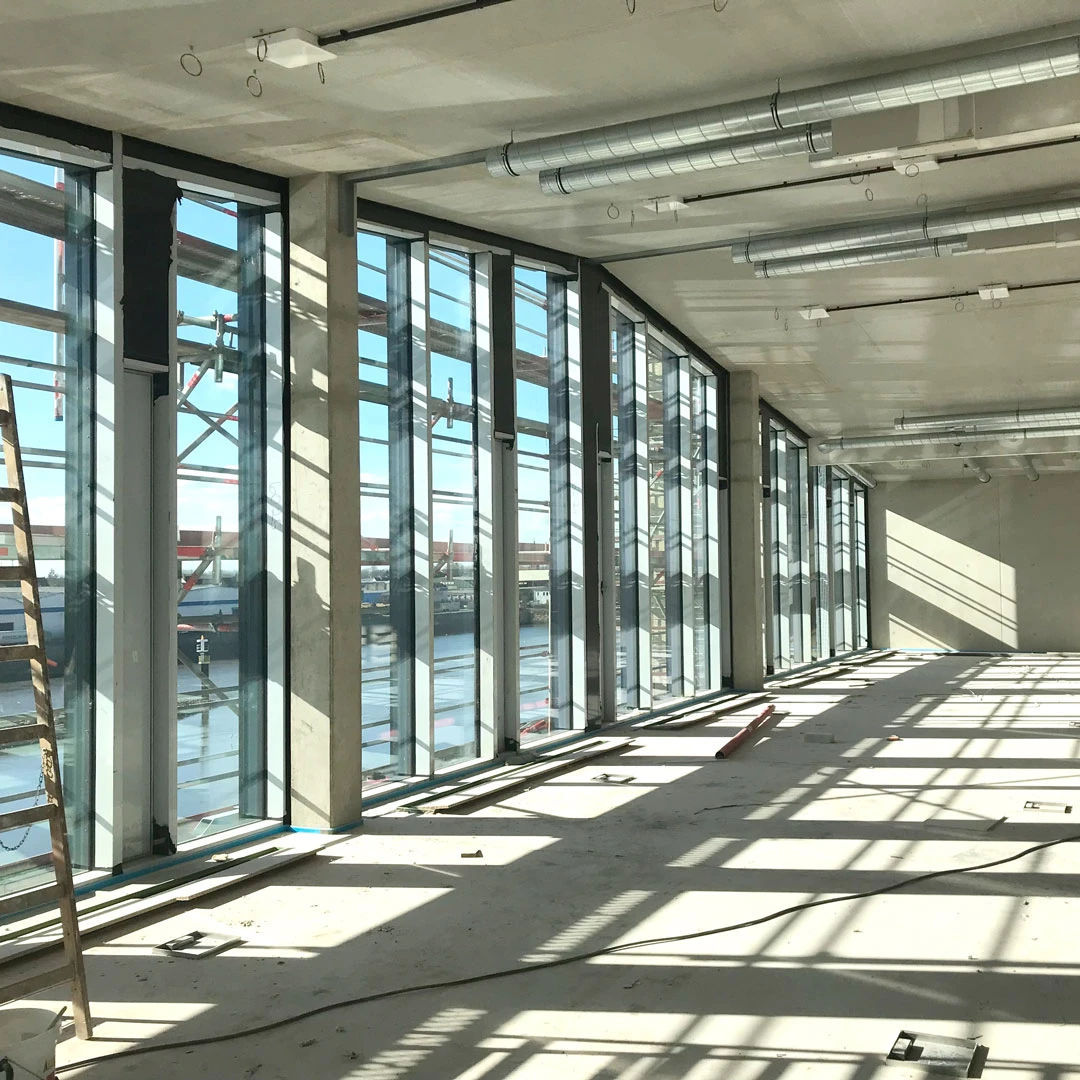 Unfinished interior with floor-to-ceiling glass windows facing a harbor; exposed ducts and construction debris.