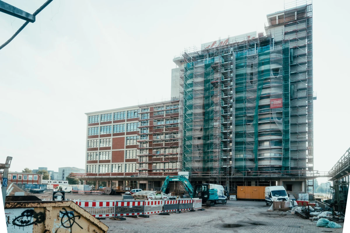 High-rise building under construction with scaffolding and green netting; foreground shows red-and-white barriers, an excavator, and parked trucks.