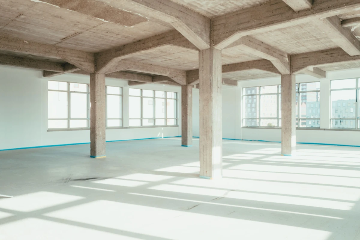 Empty unfinished commercial interior with exposed concrete columns and beams, large windows, and sunlight casting long shadows.
