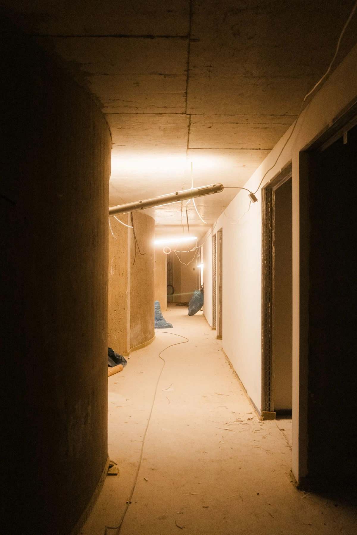 Dimly lit construction corridor with concrete walls, exposed pipes, hanging wires, and cables along a dusty floor.