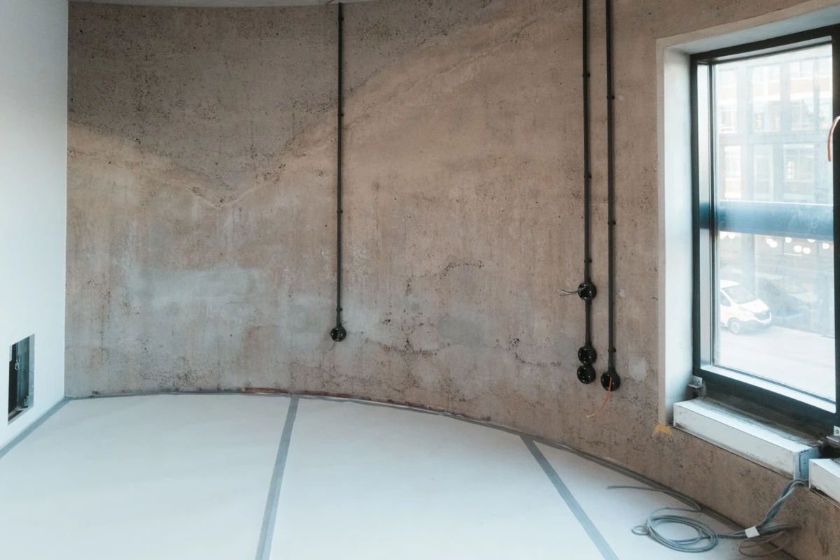Construction-site interior with a curved concrete wall, exposed electrical conduits, and a large window overlooking a city street.