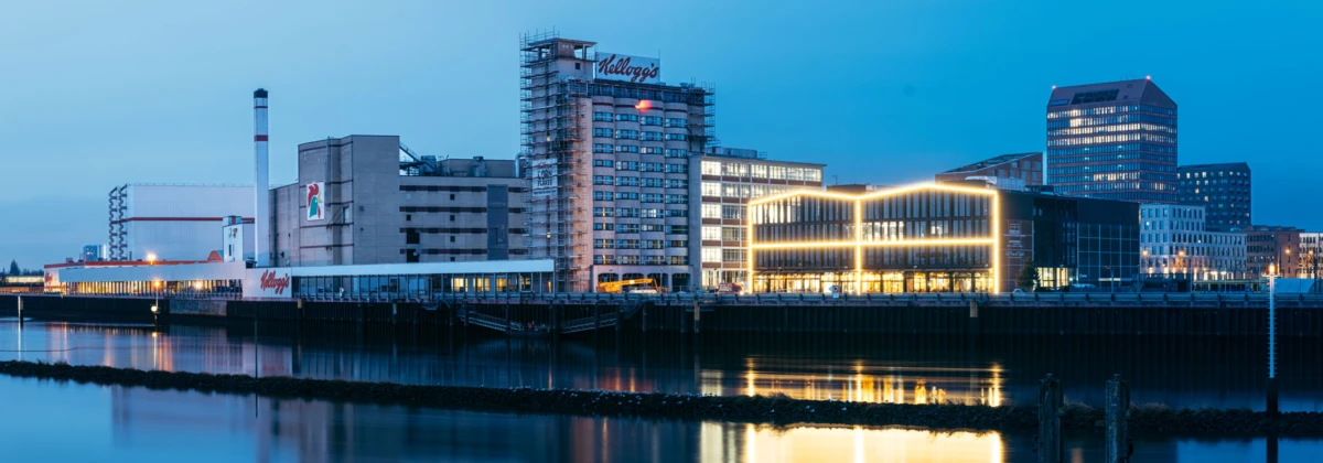 Industrial riverfront at dusk: smokestack and warehouses beside a modern, illuminated building, reflecting on calm water.