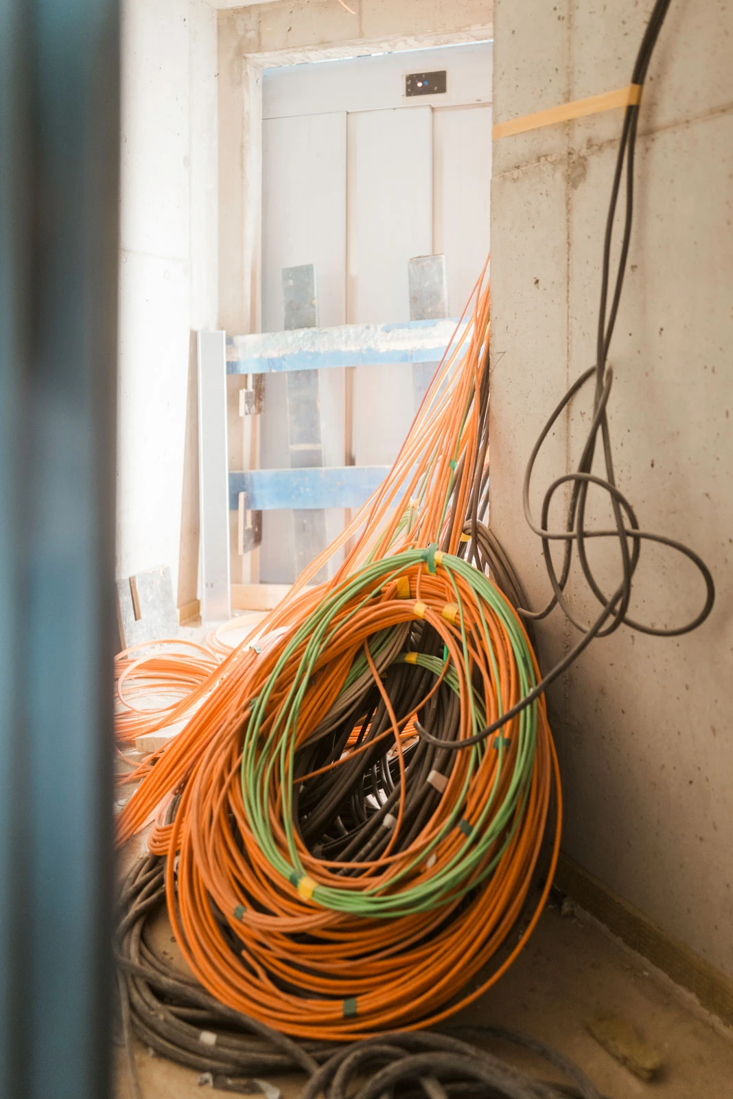 Coiled orange, green, and black electrical cables on a concrete construction site floor near an unfinished wall and elevator door.