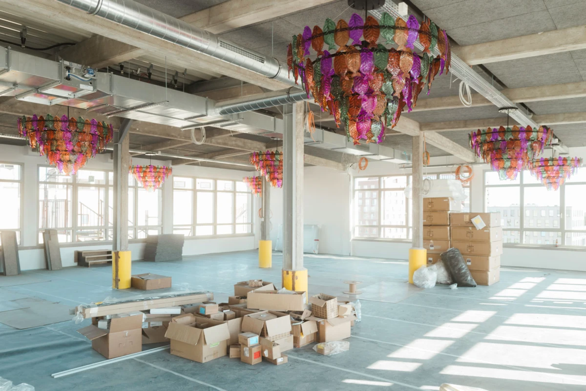 Open industrial loft under renovation, decorating with multicolored hanging lantern garlands; scattered cardboard boxes and packing materials on a blue floor.
