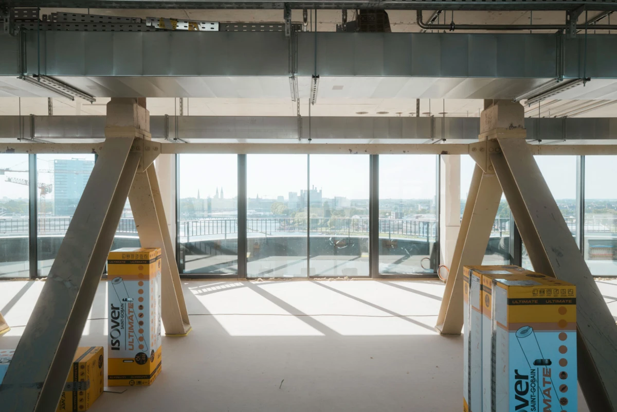Unfinished industrial interior with angled concrete columns and steel beams; boxes on the floor, city skyline visible through large windows.
