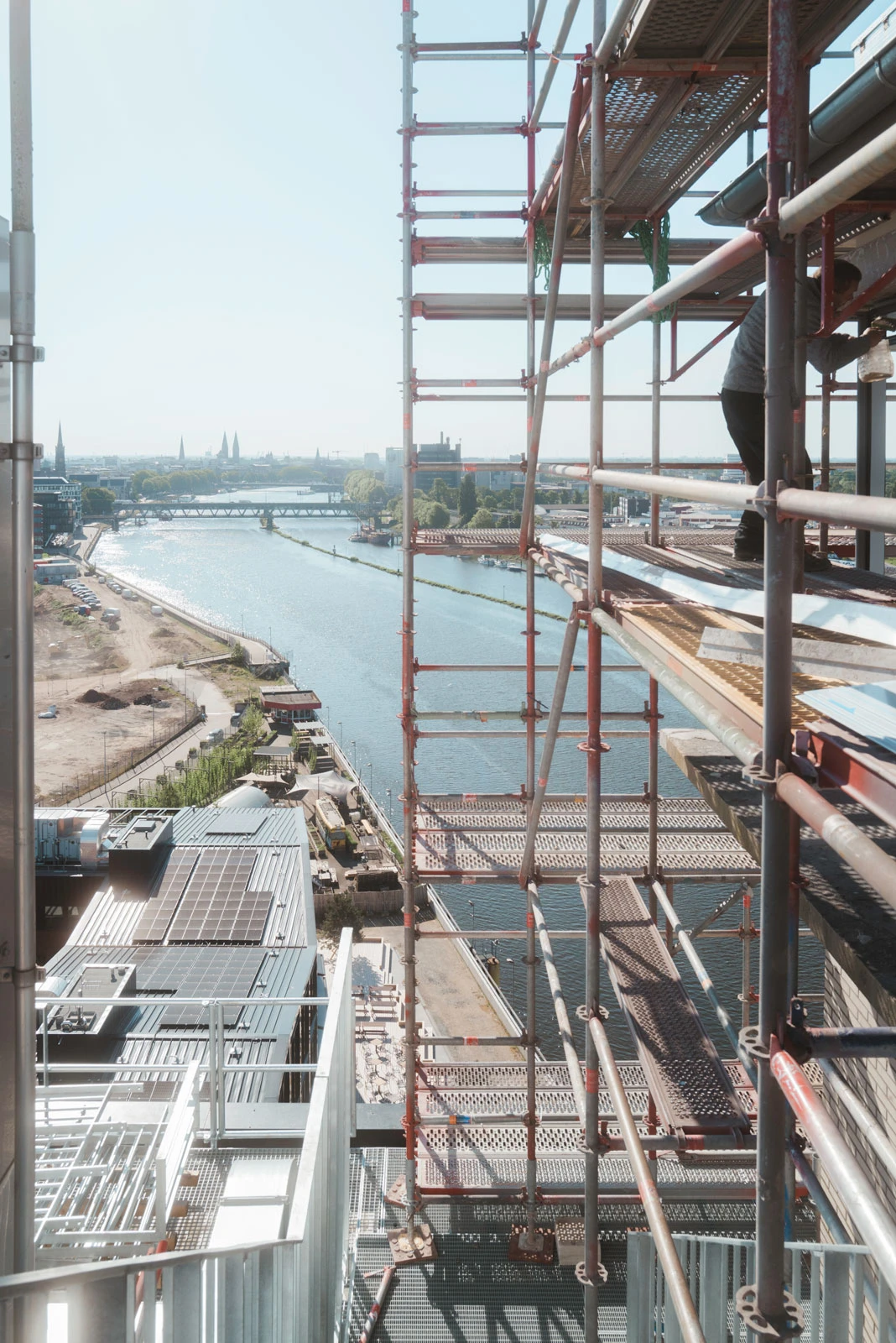 Construction worker standing on metal scaffolding attached to a high-rise overlooking a river and city skyline.