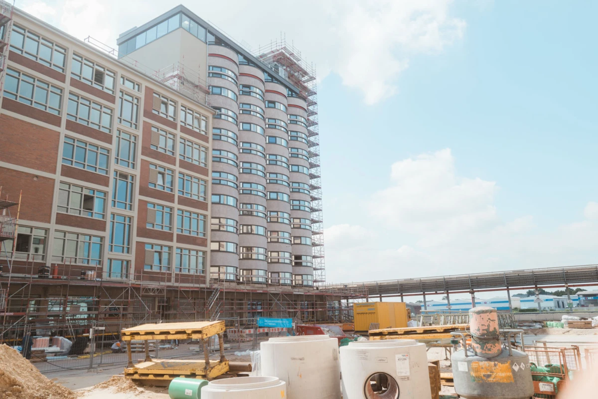 Brick-and-glass mid-rise building under construction with scaffolding; pallets and pipes in the foreground at an outdoor site.