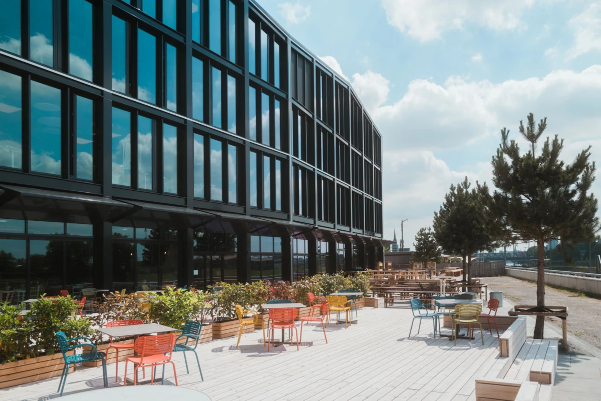 Glass-fronted office building with an outdoor terrace, colorful metal chairs and tables, and potted plants under a blue sky.