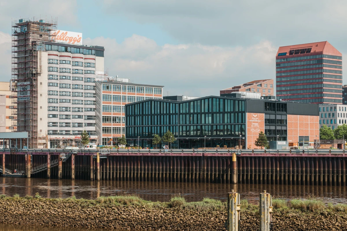 Glass-fronted office building along a riverfront with brick and dark high-rise structures in a sunny cityscape.